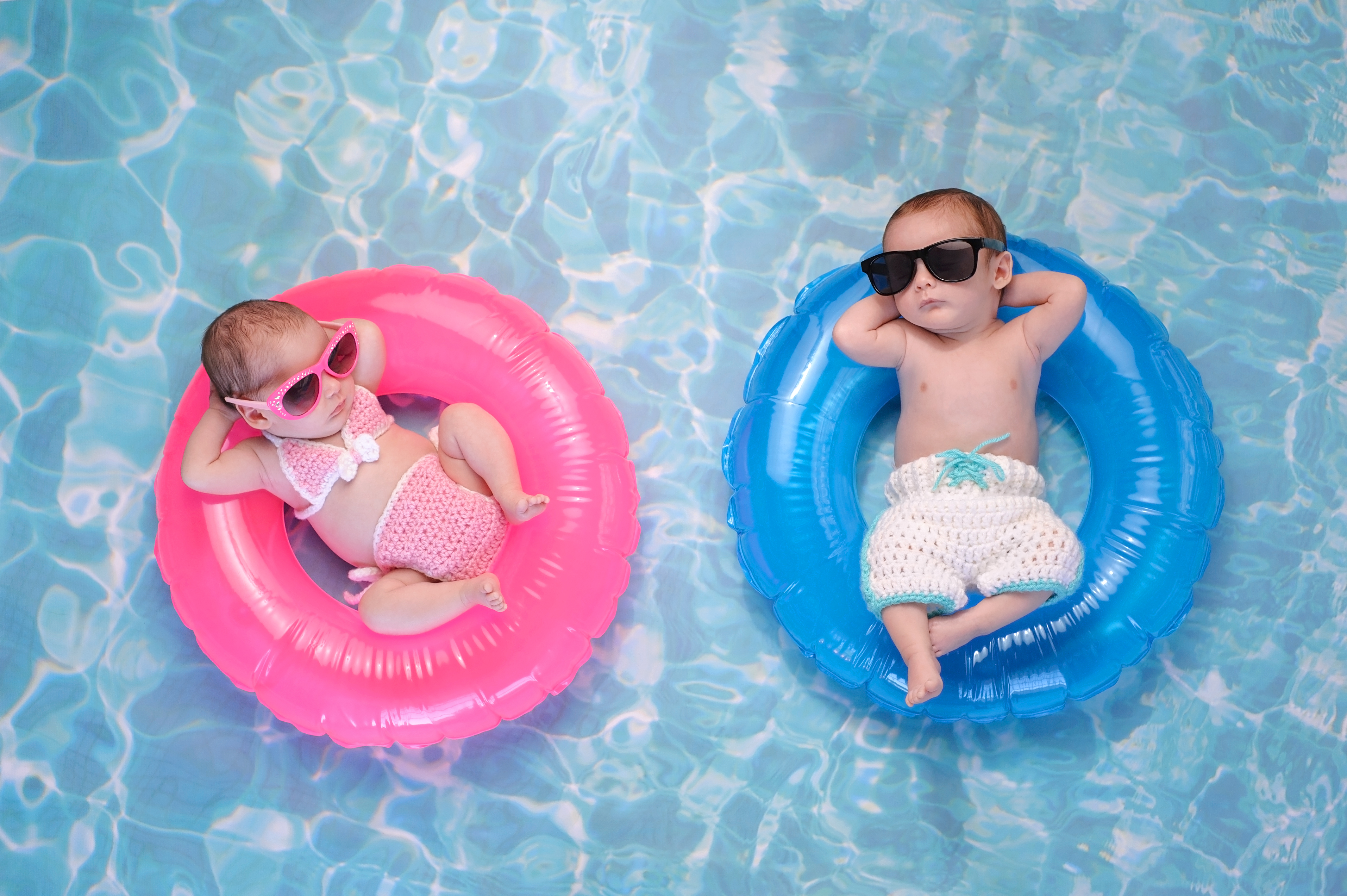 Two month old twin baby sister and brother sleeping on tiny, inflatable, pink and blue swim rings. They are wearing crocheted swimsuits and sunglasses.